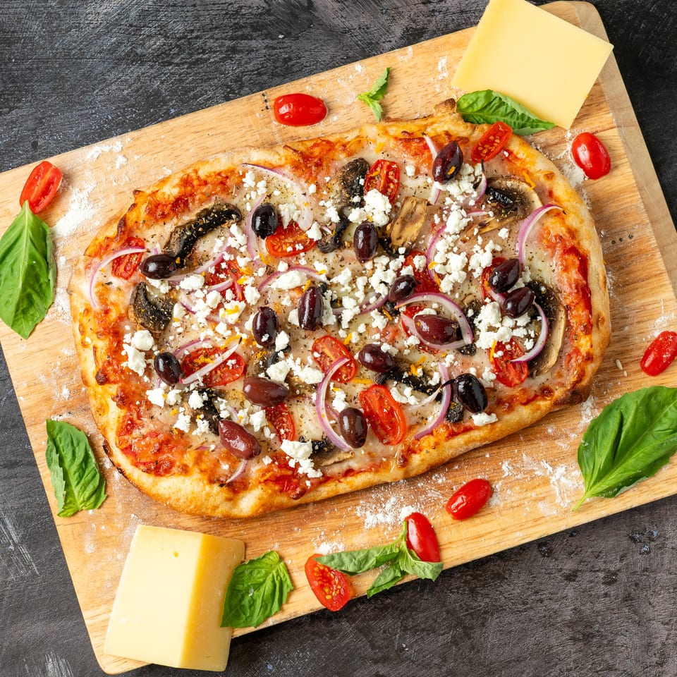 Greek-style pizza topped with olives, feta, mushrooms, tomatoes, and red onion on a wooden board with basil leaves.