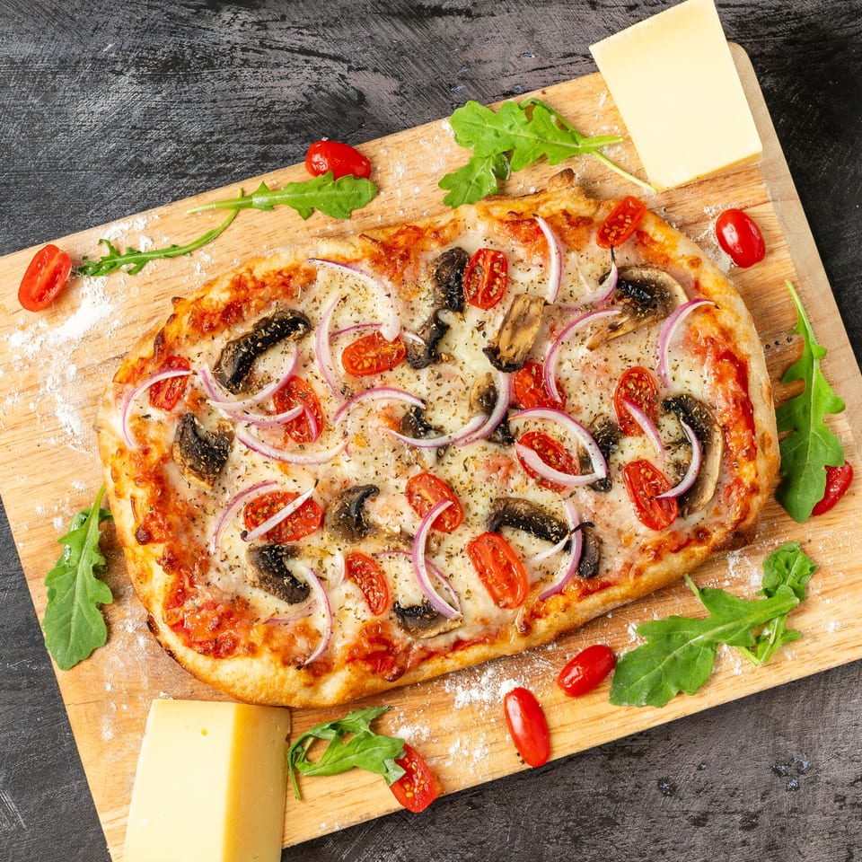 Overhead view of a vegetable pizza topped with mushrooms, tomatoes, red onion, and herbs on a wooden board.