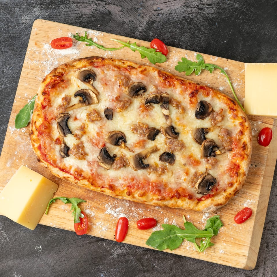 Cheesy mushroom and sausage pizza on a floured wooden board, garnished with arugula, cherry tomatoes, and cheese.