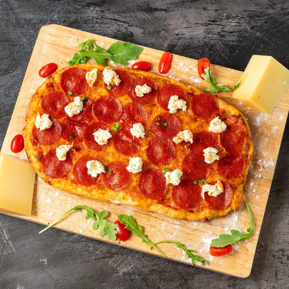 Pepperoni pizza topped with dollops of soft cheese, arugula, cherry tomatoes, and cheese wedges on a wooden board.